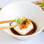 Japanese style hot tofu yudofu served in a soy sauce dashi broth being picked up with a pair of chopsticks.