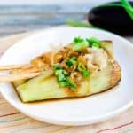 Japanese roasted eggplant yakinasu served on a white plate topped with bonito flakes, green onions, and dashi soy sauce.
