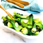 A stir fry bok choy served on a plate topped with toasted sesame seeds being picked up with a pair of chopsticks.