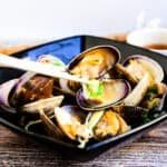 Japanese style steamed clams in a Japanese ginger sake broth served in a black bowl with chopped green onions being picked up with a pair of chopsticks.
