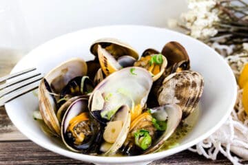 Steamed Manila clams in a white wine broth topped with chopped green onions served in a white bowl with a sliver fork.