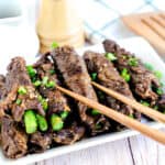 Japanese style soy glazed asparagus beef roll ups served on a white plate topped with Japanese chill powder being picked up with a pair of chopsticks.