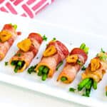 Bacon wrapped asparagus appetizers served on a white plate topped with garlic chips and Sriracha.