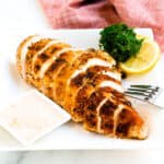 Sliced baked spicy chicken breast served on a plate topped with slices of lemon and fresh parsley served with a spicy garlic mayo dipping sauce.
