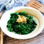 Japanese style spinach salad topped with bonito flakes served in a white bowl with a pair of chopsticks.