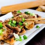 Pan fried ginger pork belly served on a white plate topped with green onions being picked up with a pair of chopsticks.