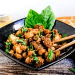 Asian style popcorn fried chicken served in a black bowl topped with fresh chopped basil being picked up with chopsticks.