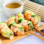 Lobster sushi roll served on a wooden sushi plate being picked up with a pair of chopsticks.