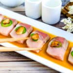 Japanese yellowtail carpaccio sashimi served on a white plate topped with sliced jalapeno and fried garlic chips being picked up with a pair of chopsticks.