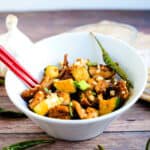 Kung pao chicken severed in a white bowl topped with green onions, chopped macadamia nuts and green pepper.