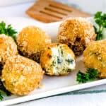 Deep fried spinach and artichoke cream cheese balls showing cross sectional cut served on a white plate.